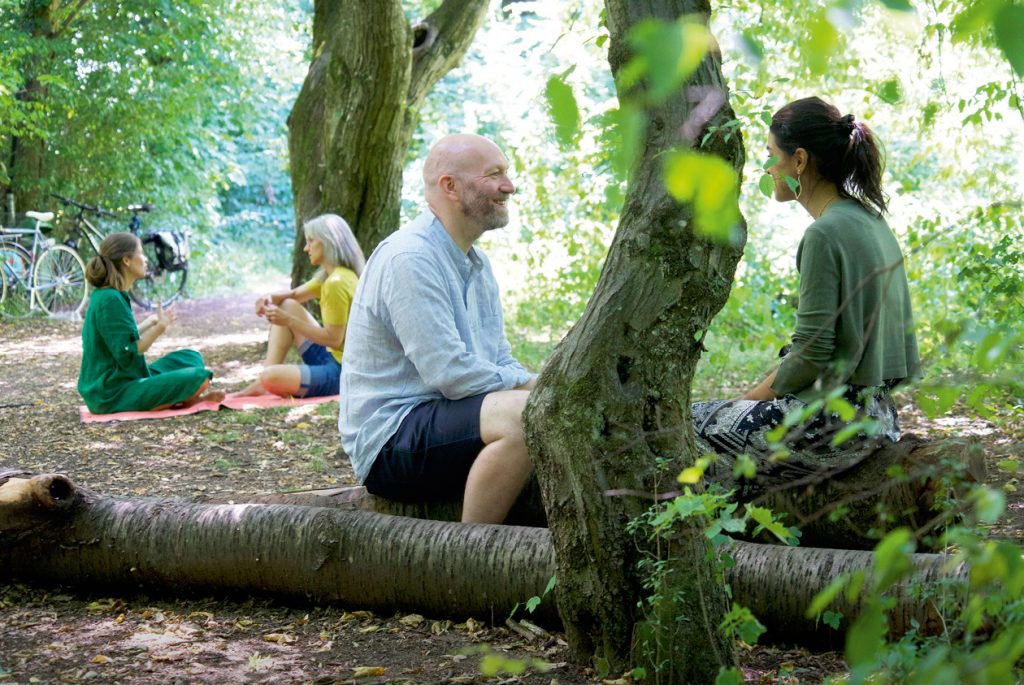 Zwei Personen sitzen auf einem Baumstamm und sehen sich an. Im Hintergrund sitzen weitere Menschen in der Natur.